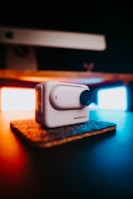 A compact, white action camera labeled 'Insta360 GO 3' sits on a small, textured mat. It is prominently featured in the foreground, with a blurred background showcasing warm and cool lighting providing contrast and ambiance.