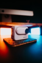 A compact, white action camera labeled 'Insta360 GO 3' sits on a small, textured mat. It is prominently featured in the foreground, with a blurred background showcasing warm and cool lighting providing contrast and ambiance.