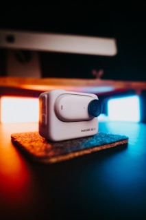 A compact, white action camera labeled 'Insta360 GO 3' sits on a small, textured mat. It is prominently featured in the foreground, with a blurred background showcasing warm and cool lighting providing contrast and ambiance.