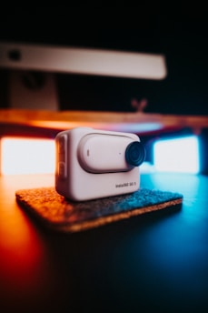 A compact, white action camera labeled 'Insta360 GO 3' sits on a small, textured mat. It is prominently featured in the foreground, with a blurred background showcasing warm and cool lighting providing contrast and ambiance.