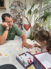 A man and a young girl are sitting at a table covered with a daisy-patterned cloth. The girl is focused on painting the man's nails with a set of colorful nail polishes. The man appears relaxed, resting his head on one hand. Behind them, a collection of family photos is displayed on a wall, along with decorative elements like a hanging heart and potted plants near a window.