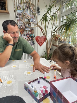 A man and a young girl are sitting at a table covered with a daisy-patterned cloth. The girl is focused on painting the man's nails with a set of colorful nail polishes. The man appears relaxed, resting his head on one hand. Behind them, a collection of family photos is displayed on a wall, along with decorative elements like a hanging heart and potted plants near a window.