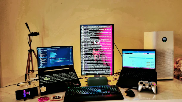 A workspace setup featuring three computer screens; two laptops and one vertically oriented monitor displaying lines of code and text with a bright pink graphic. In the foreground, a keyboard and mouse rest on the desk. To the right, a game controller is visible near another laptop. An air purifier is positioned on the far right, and a smartphone lies to the left. Additional items include a tripod and a small portable speaker on the desk.