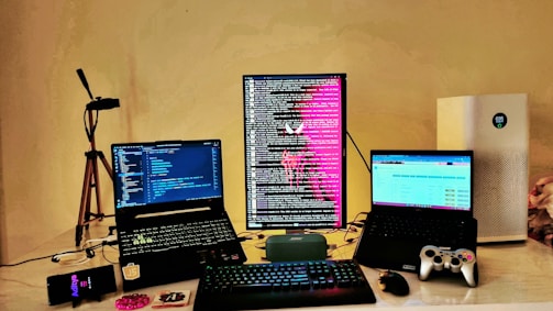 A workspace setup featuring three computer screens; two laptops and one vertically oriented monitor displaying lines of code and text with a bright pink graphic. In the foreground, a keyboard and mouse rest on the desk. To the right, a game controller is visible near another laptop. An air purifier is positioned on the far right, and a smartphone lies to the left. Additional items include a tripod and a small portable speaker on the desk.