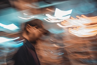 A dynamic, abstract image featuring a blurred figure in the foreground, likely a person wearing a cap. The background is a mix of swirling lights and vibrant colors, creating a sense of motion and fluidity.