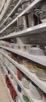 Shelves filled with a variety of kitchenware, including metal colanders, bowls, and stacks of ceramic plates in different colors and patterns.
