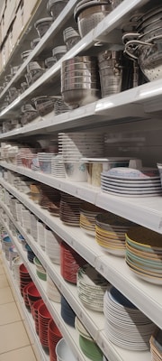 Shelves filled with a variety of kitchenware, including metal colanders, bowls, and stacks of ceramic plates in different colors and patterns.