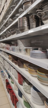 Shelves filled with a variety of kitchenware, including metal colanders, bowls, and stacks of ceramic plates in different colors and patterns.