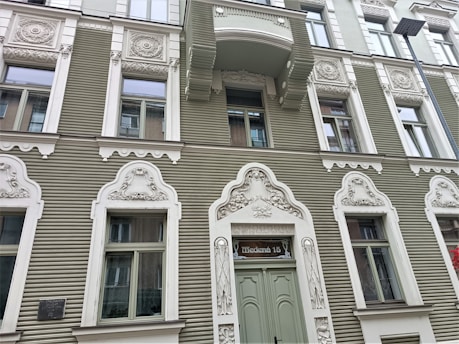 A detailed façade of a building with ornately designed white window frames and decorations on a sage green exterior. Each window is framed with intricate carvings and motifs. A sign above the main door reads 'Tērbatas 15'. There is a balcony above one of the windows, and a street lamp is visible on the right side.