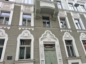 A detailed façade of a building with ornately designed white window frames and decorations on a sage green exterior. Each window is framed with intricate carvings and motifs. A sign above the main door reads 'Tērbatas 15'. There is a balcony above one of the windows, and a street lamp is visible on the right side.