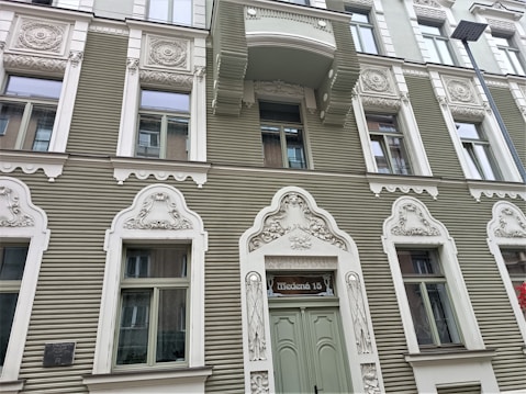A detailed fa&ccedil;ade of a building with ornately designed white window frames and decorations on a sage green exterior. Each window is framed with intricate carvings and motifs. A sign above the main door reads 'Tērbatas 15'. There is a balcony above one of the windows, and a street lamp is visible on the right side.