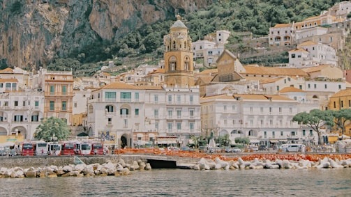 A picturesque coastal town with a blend of historic architecture and modern elements. The scene includes a prominent bell tower surrounded by numerous buildings with warm-colored roofs, all set against a backdrop of lush greenery and steep cliffs. The town is bordered by a serene body of water, in front of which are some red buses and vehicles visible near the shore alongside rocky barriers.