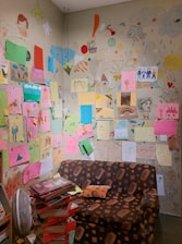 A small room with walls covered in colorful children's drawings, featuring various subjects like houses, rainbows, and people. A brown patterned couch sits against the wall, and a stack of books is piled high in the corner. The artwork is diverse, with an array of vibrant papers and sketches directly on the wall.