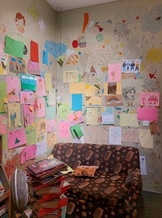 A small room with walls covered in colorful children's drawings, featuring various subjects like houses, rainbows, and people. A brown patterned couch sits against the wall, and a stack of books is piled high in the corner. The artwork is diverse, with an array of vibrant papers and sketches directly on the wall.