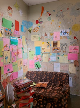 A small room with walls covered in colorful children's drawings, featuring various subjects like houses, rainbows, and people. A brown patterned couch sits against the wall, and a stack of books is piled high in the corner. The artwork is diverse, with an array of vibrant papers and sketches directly on the wall.