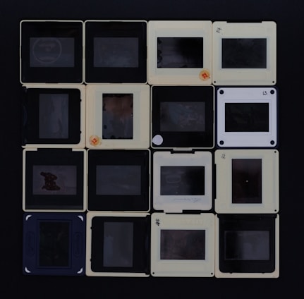 A collection of sixteen slide film mounts, arranged in a four-by-four grid. The mounts are mostly beige and black, with some showing faded writing or labels. Each slide has a transparent center where the film is located, though the images on the film are not clearly visible.