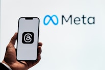 A person holds a smartphone displaying a logo with an '@' symbol inside a square. In the background, the logo and name 'Meta' are visible, with the infinity symbol in blue next to the word 'Meta' in black.