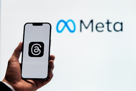 A person holds a smartphone displaying a logo with an '@' symbol inside a square. In the background, the logo and name 'Meta' are visible, with the infinity symbol in blue next to the word 'Meta' in black.