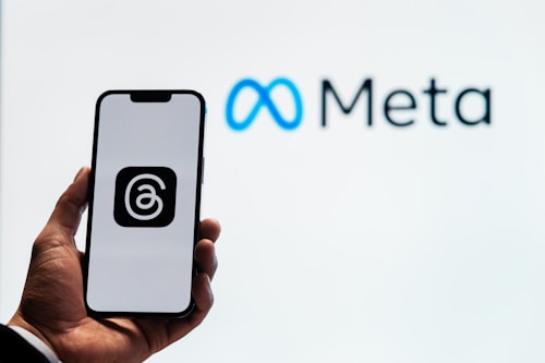 A person holds a smartphone displaying a logo with an '@' symbol inside a square. In the background, the logo and name 'Meta' are visible, with the infinity symbol in blue next to the word 'Meta' in black.
