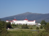 A large, historic hotel with red roofs sits amidst lush greenery, surrounded by vast woodlands and a mountainous backdrop. The building is multi-storied and painted white, with numerous windows and architectural features. A clear blue sky complements the scene, and a winding road leads up to the entrance of the hotel.