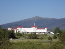 A large, historic hotel with red roofs sits amidst lush greenery, surrounded by vast woodlands and a mountainous backdrop. The building is multi-storied and painted white, with numerous windows and architectural features. A clear blue sky complements the scene, and a winding road leads up to the entrance of the hotel.