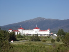 A large, historic hotel with red roofs sits amidst lush greenery, surrounded by vast woodlands and a mountainous backdrop. The building is multi-storied and painted white, with numerous windows and architectural features. A clear blue sky complements the scene, and a winding road leads up to the entrance of the hotel.