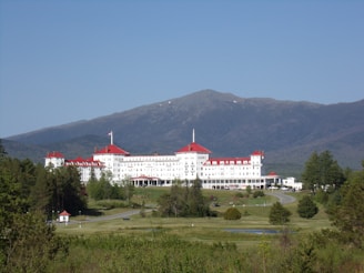 A large, historic hotel with red roofs sits amidst lush greenery, surrounded by vast woodlands and a mountainous backdrop. The building is multi-storied and painted white, with numerous windows and architectural features. A clear blue sky complements the scene, and a winding road leads up to the entrance of the hotel.