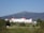 A large, historic hotel with red roofs sits amidst lush greenery, surrounded by vast woodlands and a mountainous backdrop. The building is multi-storied and painted white, with numerous windows and architectural features. A clear blue sky complements the scene, and a winding road leads up to the entrance of the hotel.