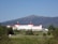 A large, historic hotel with red roofs sits amidst lush greenery, surrounded by vast woodlands and a mountainous backdrop. The building is multi-storied and painted white, with numerous windows and architectural features. A clear blue sky complements the scene, and a winding road leads up to the entrance of the hotel.