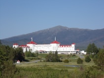 A large, historic hotel with red roofs sits amidst lush greenery, surrounded by vast woodlands and a mountainous backdrop. The building is multi-storied and painted white, with numerous windows and architectural features. A clear blue sky complements the scene, and a winding road leads up to the entrance of the hotel.
