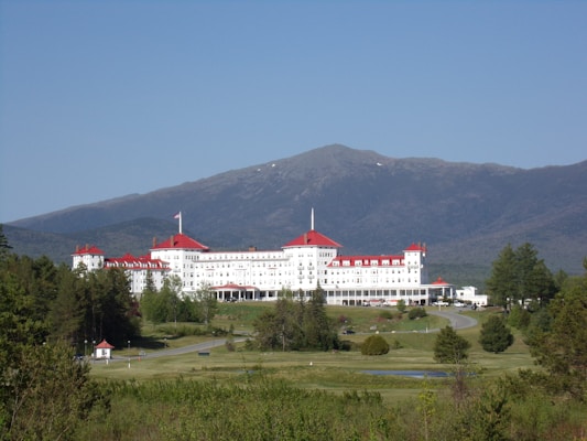 A large, historic hotel with red roofs sits amidst lush greenery, surrounded by vast woodlands and a mountainous backdrop. The building is multi-storied and painted white, with numerous windows and architectural features. A clear blue sky complements the scene, and a winding road leads up to the entrance of the hotel.