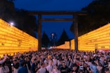 A large crowd of people is gathered at a traditional festival or event during the evening. The scene is framed by two long walls of illuminated yellow lanterns with writing on them, and a large torii gate stands in the center. In the background, there are tall trees and a modern building further away. The sky is deep blue, suggesting late in the day.