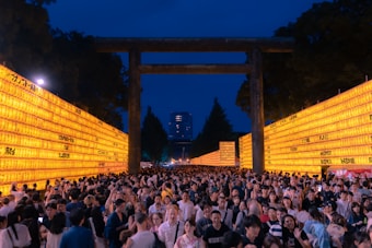A large crowd of people is gathered at a traditional festival or event during the evening. The scene is framed by two long walls of illuminated yellow lanterns with writing on them, and a large torii gate stands in the center. In the background, there are tall trees and a modern building further away. The sky is deep blue, suggesting late in the day.