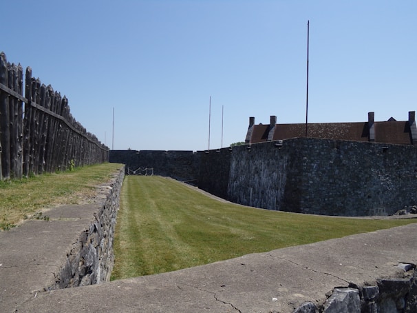 A stone fortress with tall walls and a neatly trimmed grassy area inside. Wooden barricades line one side, and there is a clear blue sky above.