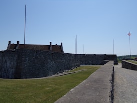 A stone fortification with tall bastions and a sloped grass embankment appears under a clear blue sky. Flagpoles are visible, one with an American flag. The structure suggests a historical military purpose, surrounded by a path and lush greenery.