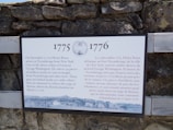 An information plaque is mounted on a stone wall. The plaque details historical events between 1775 and 1776 concerning Henry Knox and his transportation of artillery from Ticonderoga to assist George Washington during the American Revolutionary War. The text is written in both English and French.