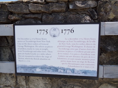 An information plaque is mounted on a stone wall. The plaque details historical events between 1775 and 1776 concerning Henry Knox and his transportation of artillery from Ticonderoga to assist George Washington during the American Revolutionary War. The text is written in both English and French.