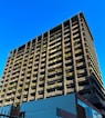 A towering building with a modern architectural design rises against a clear blue sky. The structure features numerous balconies adorned with greenery, giving it an eco-friendly appearance. The bottom of the building showcases commercial signage, hinting at a mixed-use purpose. The sunlight casts shadows on one side, highlighting the grid-like structure.