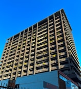 A towering building with a modern architectural design rises against a clear blue sky. The structure features numerous balconies adorned with greenery, giving it an eco-friendly appearance. The bottom of the building showcases commercial signage, hinting at a mixed-use purpose. The sunlight casts shadows on one side, highlighting the grid-like structure.