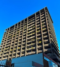 A towering building with a modern architectural design rises against a clear blue sky. The structure features numerous balconies adorned with greenery, giving it an eco-friendly appearance. The bottom of the building showcases commercial signage, hinting at a mixed-use purpose. The sunlight casts shadows on one side, highlighting the grid-like structure.