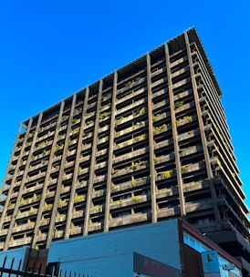 A towering building with a modern architectural design rises against a clear blue sky. The structure features numerous balconies adorned with greenery, giving it an eco-friendly appearance. The bottom of the building showcases commercial signage, hinting at a mixed-use purpose. The sunlight casts shadows on one side, highlighting the grid-like structure.