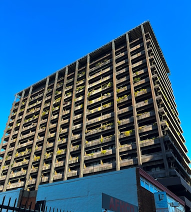 A towering building with a modern architectural design rises against a clear blue sky. The structure features numerous balconies adorned with greenery, giving it an eco-friendly appearance. The bottom of the building showcases commercial signage, hinting at a mixed-use purpose. The sunlight casts shadows on one side, highlighting the grid-like structure.