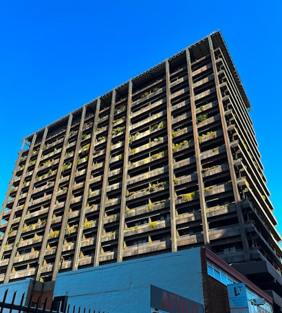 A towering building with a modern architectural design rises against a clear blue sky. The structure features numerous balconies adorned with greenery, giving it an eco-friendly appearance. The bottom of the building showcases commercial signage, hinting at a mixed-use purpose. The sunlight casts shadows on one side, highlighting the grid-like structure.