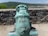 A historic bronze cannon is prominently displayed on a stone platform. The cannon features intricate carvings and designs, including a face motif on its front. The background includes a solid stone wall and a scenic view of lush green hills under a clear blue sky.