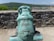A historic bronze cannon is prominently displayed on a stone platform. The cannon features intricate carvings and designs, including a face motif on its front. The background includes a solid stone wall and a scenic view of lush green hills under a clear blue sky.