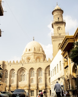 A historic mosque with intricate architecture featuring a large dome and a tall minaret. The façade is adorned with ornate designs and lattice windows. People walk on the street in front of the mosque, and cars are parked along the side. Adjacent buildings display a mix of traditional and modern elements.