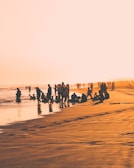 A group of diverse women, men, and children laughing together on a beach at golden hour.