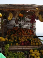 A market stall displays a variety of fresh fruits, including bananas, oranges, lemons, grapes, and tomatoes. The fruits are arranged in baskets made of woven bamboo, creating a rustic and inviting appearance. The fruits appear vibrant under the daylight, with leaves adding a natural touch.