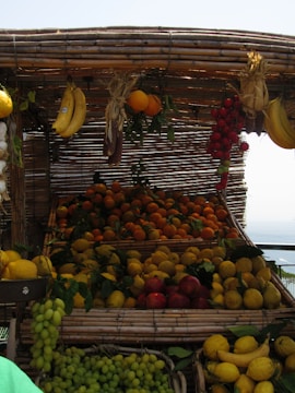 A market stall displays a variety of fresh fruits, including bananas, oranges, lemons, grapes, and tomatoes. The fruits are arranged in baskets made of woven bamboo, creating a rustic and inviting appearance. The fruits appear vibrant under the daylight, with leaves adding a natural touch.