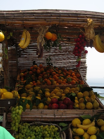 A market stall displays a variety of fresh fruits, including bananas, oranges, lemons, grapes, and tomatoes. The fruits are arranged in baskets made of woven bamboo, creating a rustic and inviting appearance. The fruits appear vibrant under the daylight, with leaves adding a natural touch.